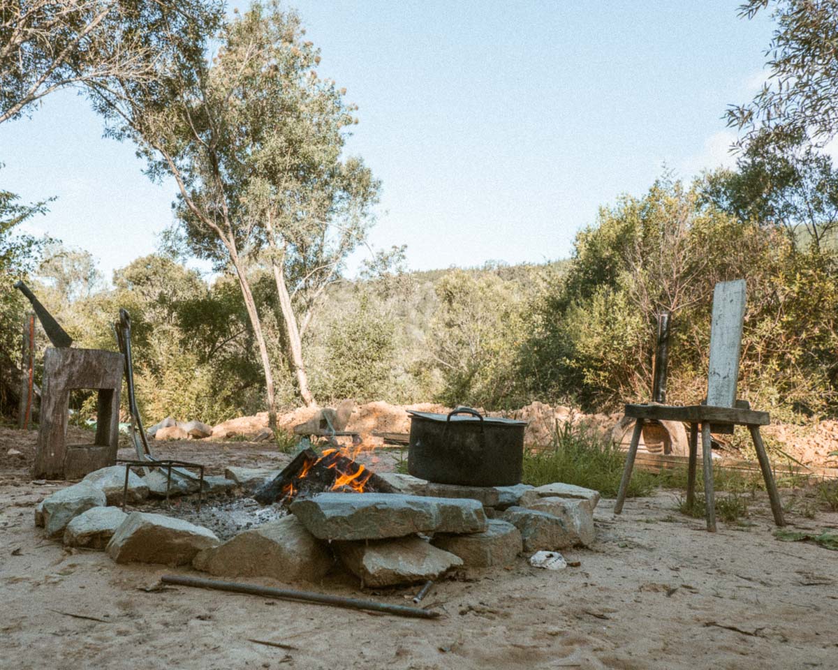Outdoor fire scene with a stone ring, visible flames, a dark metal pot, trees, and a sandy ground in a natural wooded setting.
