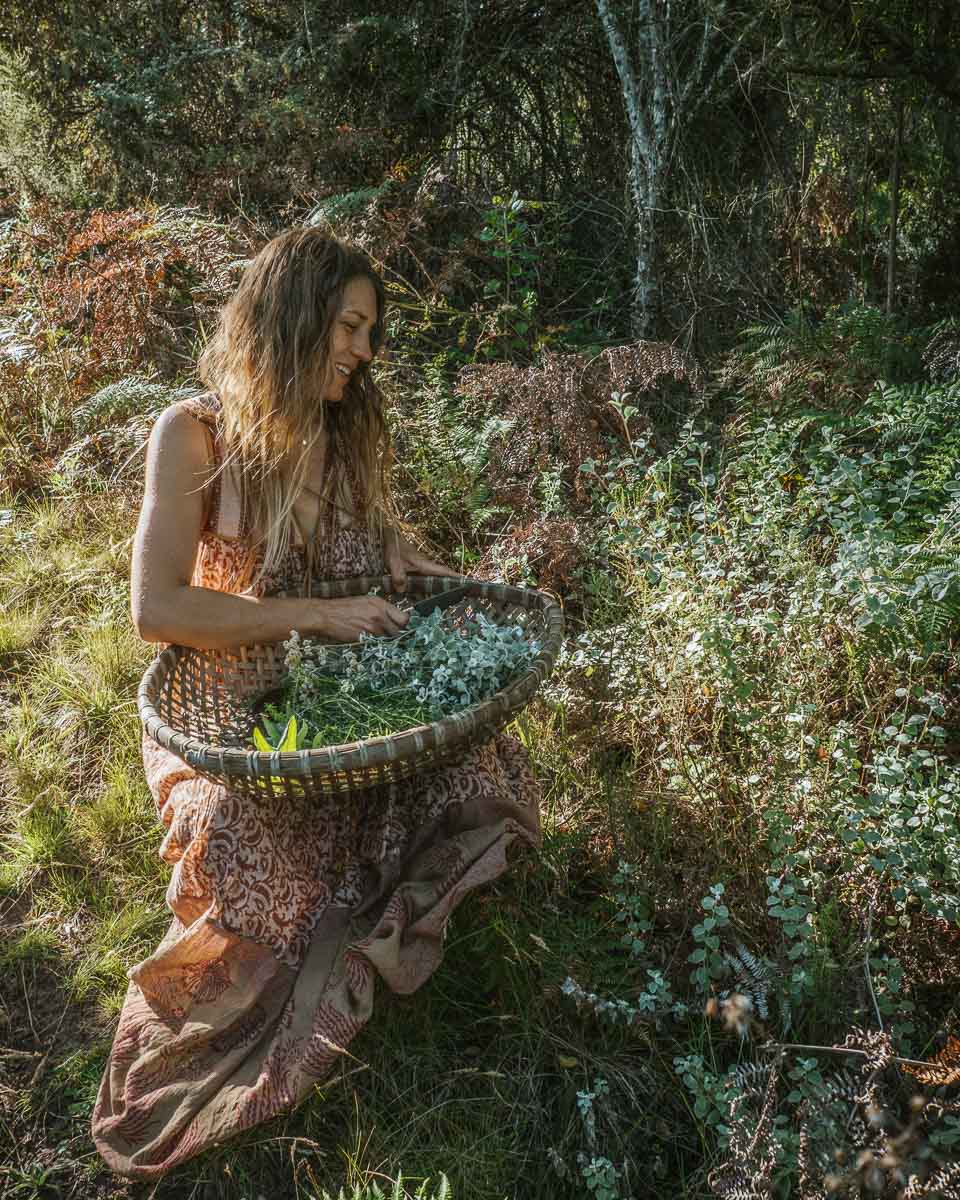 A woman in a brown, patterned dress kneels in a forest clearing, holding a large woven basket filled with green leaves and small white flowers.