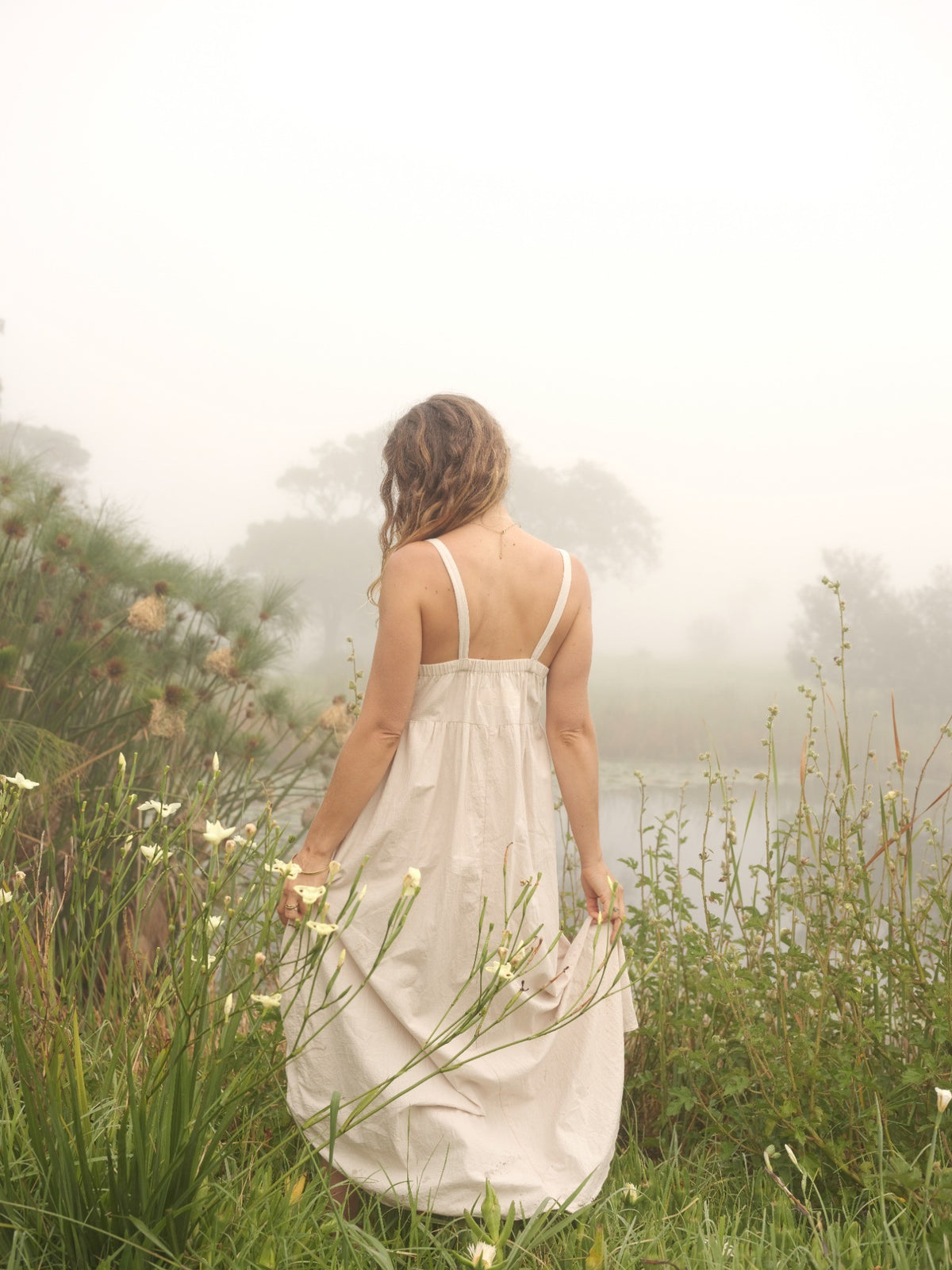 A woman stands with her back to the camera in a misty outdoor field beside water, wearing a loose white sleeveless dress.