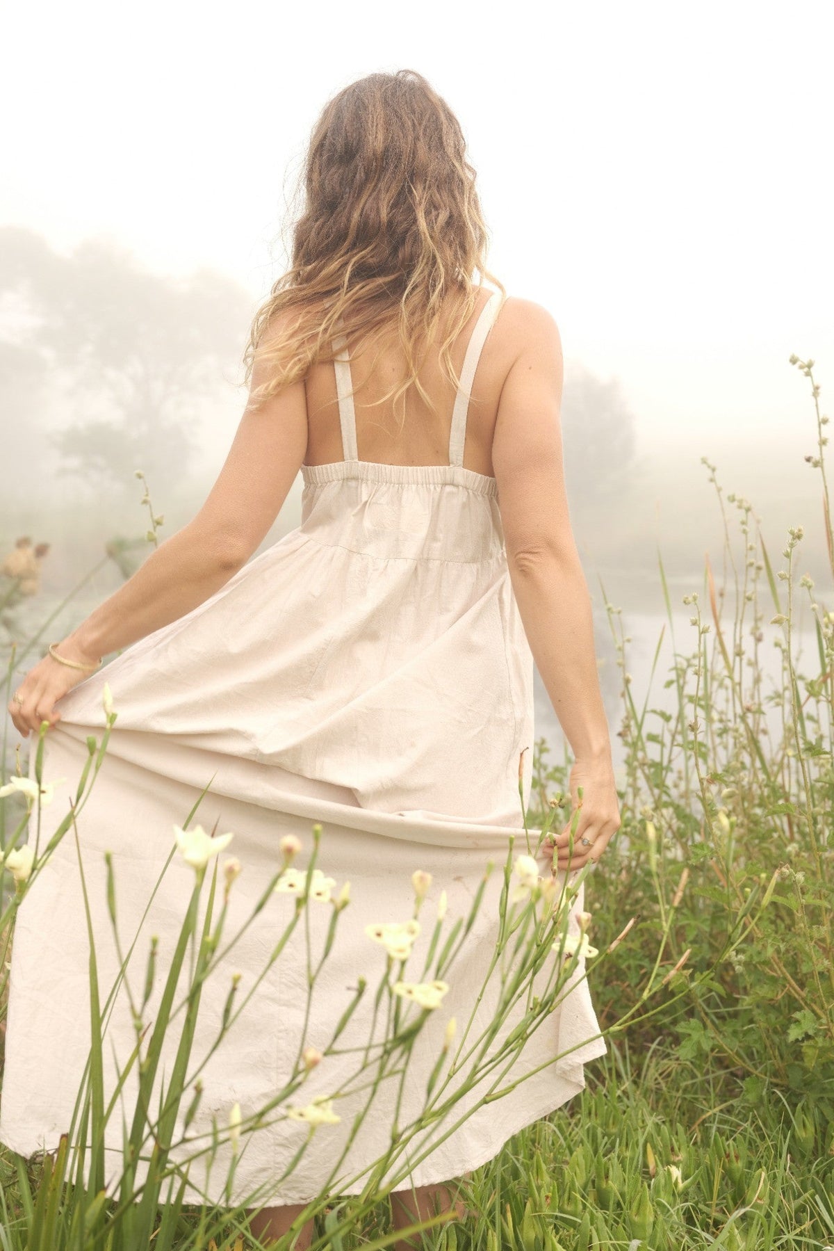 A woman stands outdoors in a misty field, viewed from behind, wearing a light beige linen slip dress and holding out the skirt to show its flow. Tall green grasses and small white wildflowers surround her.