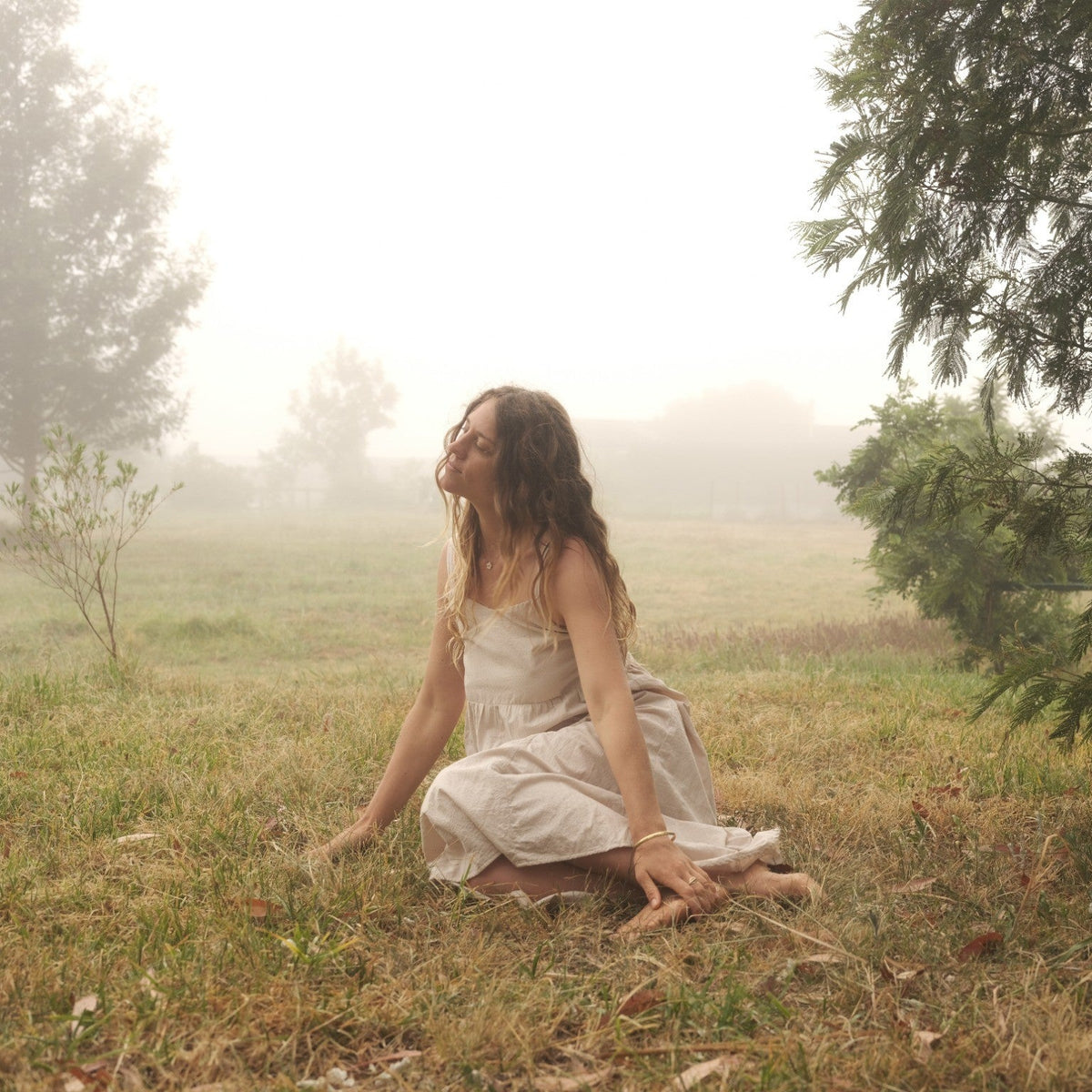 A woman in a light cream-colored sleeveless dress kneels on grass in a misty outdoor field, looking upward near trees.