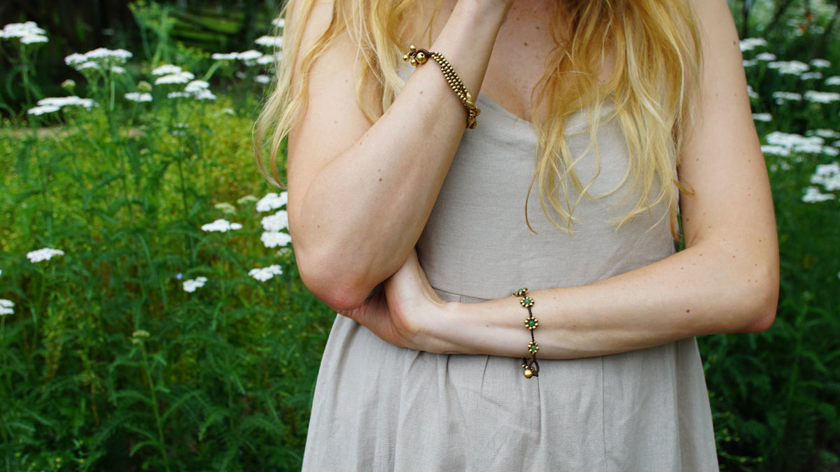 A woman outdoors in a field of green plants and white flowers, wearing a light beige dress and two gold-toned beaded bracelets.