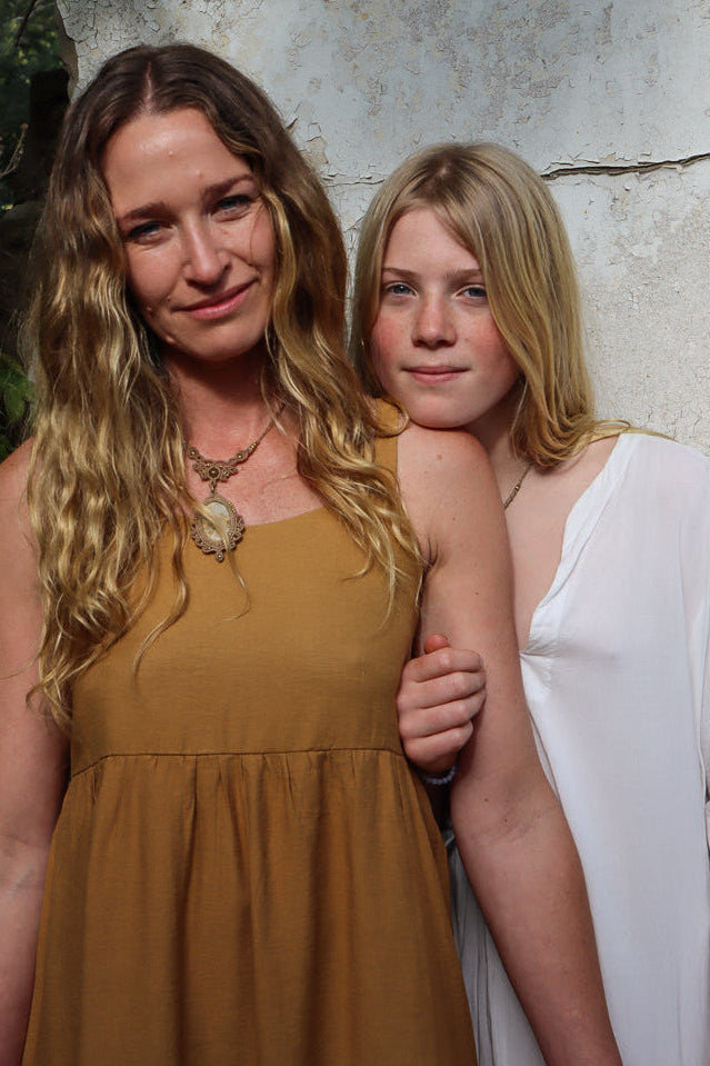Two women posing together outdoors against a textured light grey wall. The woman on the left wears a sleeveless mustard-brown dress and a large pendant necklace, while the woman on the right wears a white long-sleeve top.