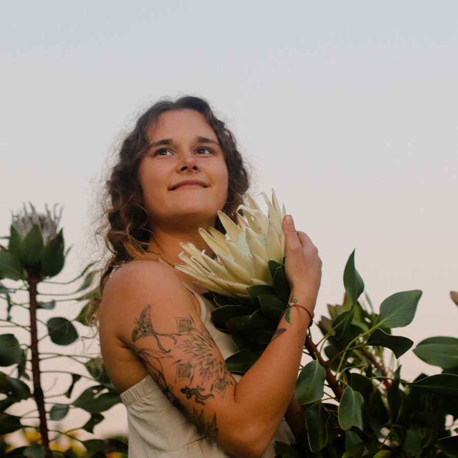 A woman with curly brown hair and arm tattoos holds a large white protea flower against a soft sky background with green leaves around her.