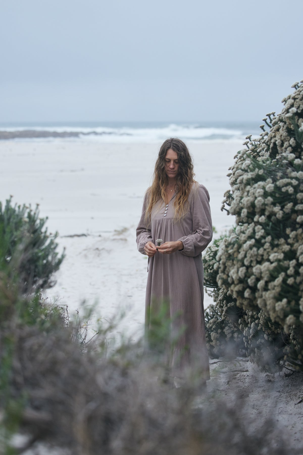 A woman wearing a long-sleeved, button-front maxi dress in a muted taupe-brown stands on a sandy beach path, framed by green and white flowering bushes under an overcast sky.