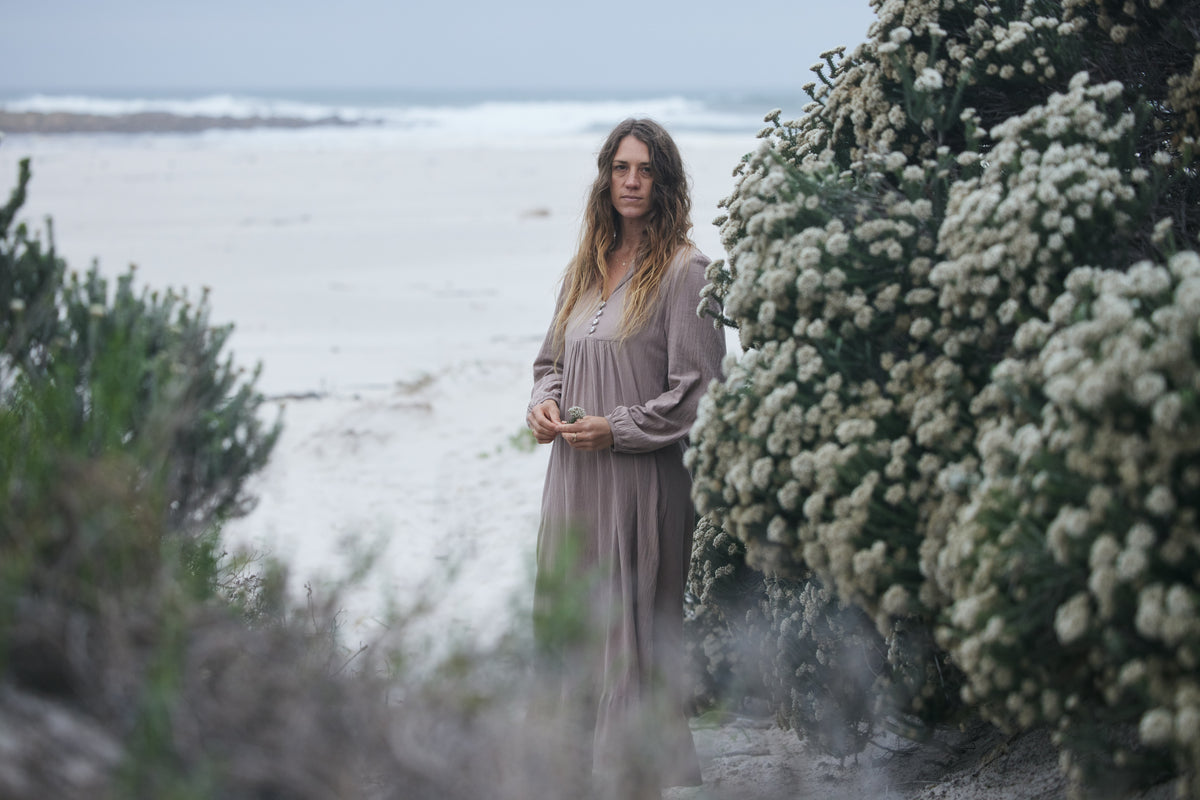 A woman stands outdoors on a beach path between flowering bushes, wearing a long, loose taupe dress. The background shows a soft-focus shoreline and ocean under an overcast sky.