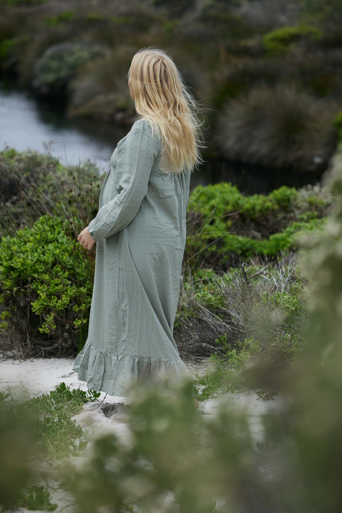 A woman with long blonde hair stands outdoors on a sandy path surrounded by green shrubs, wearing a long sage green maxi dress and looking away from the camera.