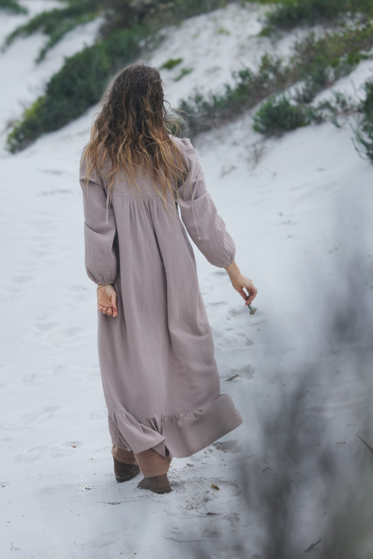 A woman wearing a long, loose-fitting dusty mauve maxi dress walks along a sandy dune path with sparse green vegetation in the background.