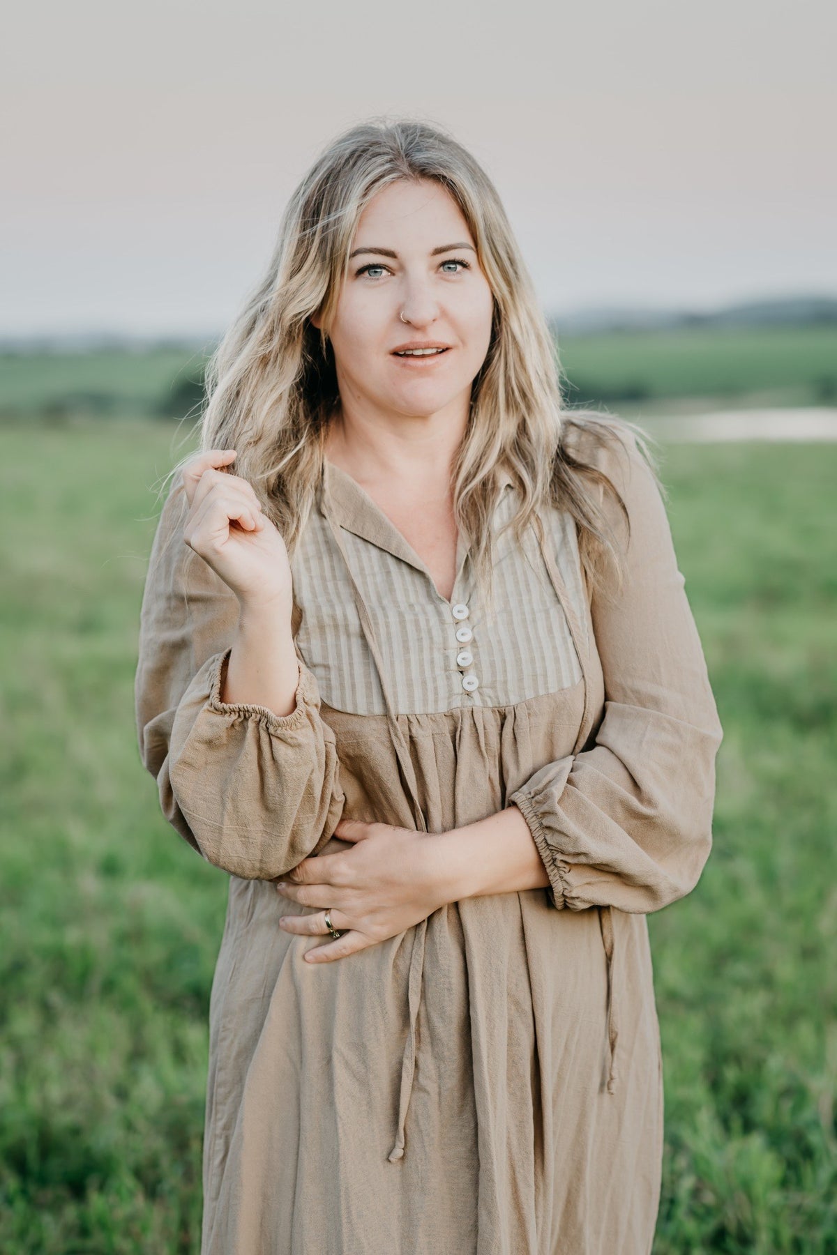 A woman with blonde hair stands in a grassy field wearing a beige and brown striped long-sleeve dress with a buttoned chest and gathered cuffs.
