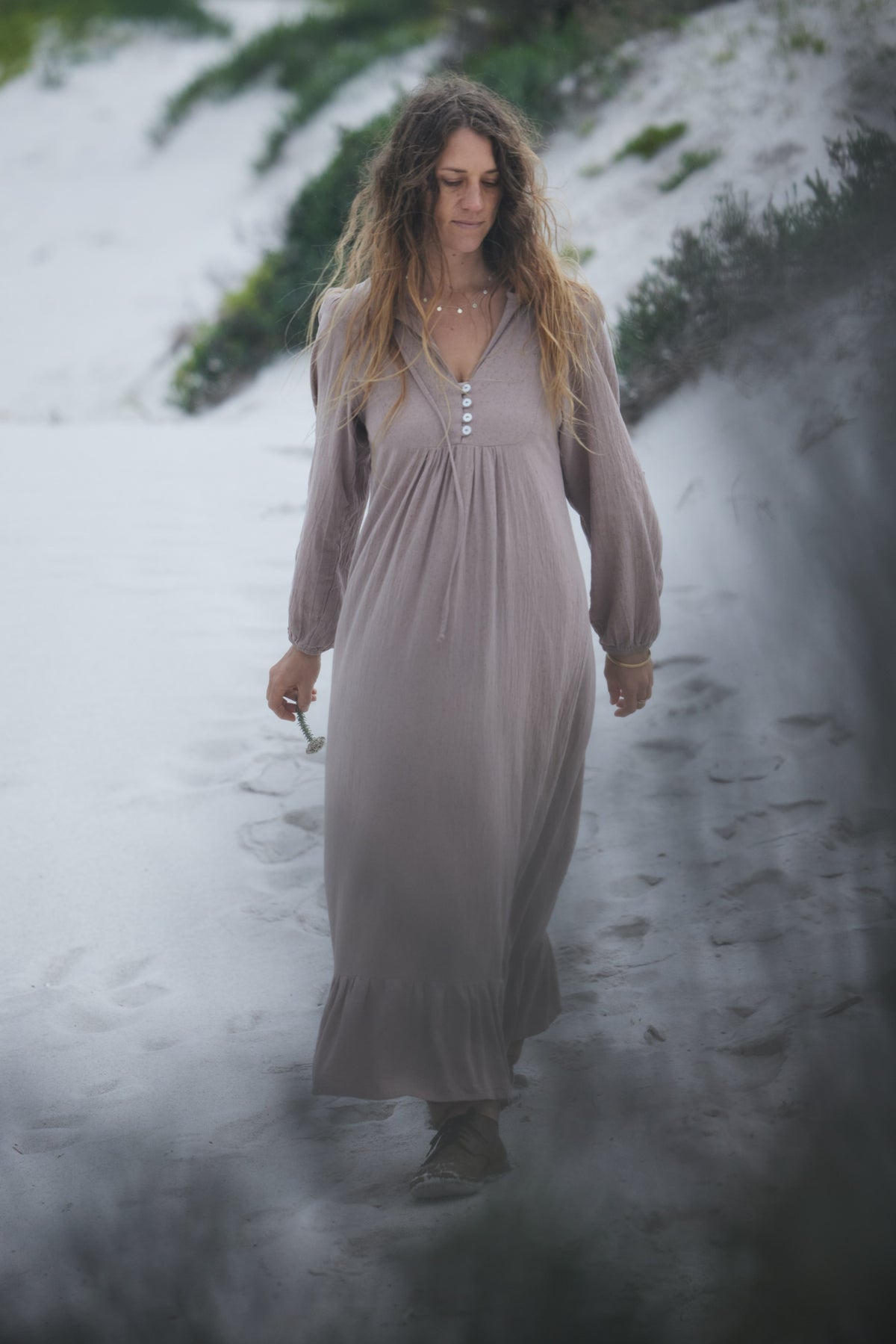 A woman wearing a long, loose-fitting taupe maxi dress walks along a sandy path surrounded by mist and dune vegetation.