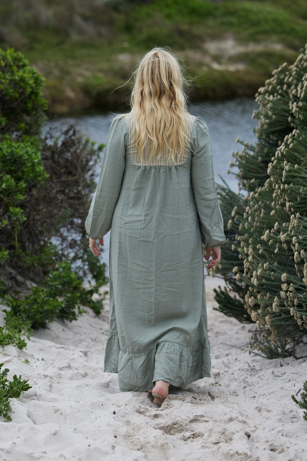 A woman with long blonde hair is seen from behind, wearing a sage green linen maxi dress while walking barefoot on a sandy path surrounded by green plants and water.