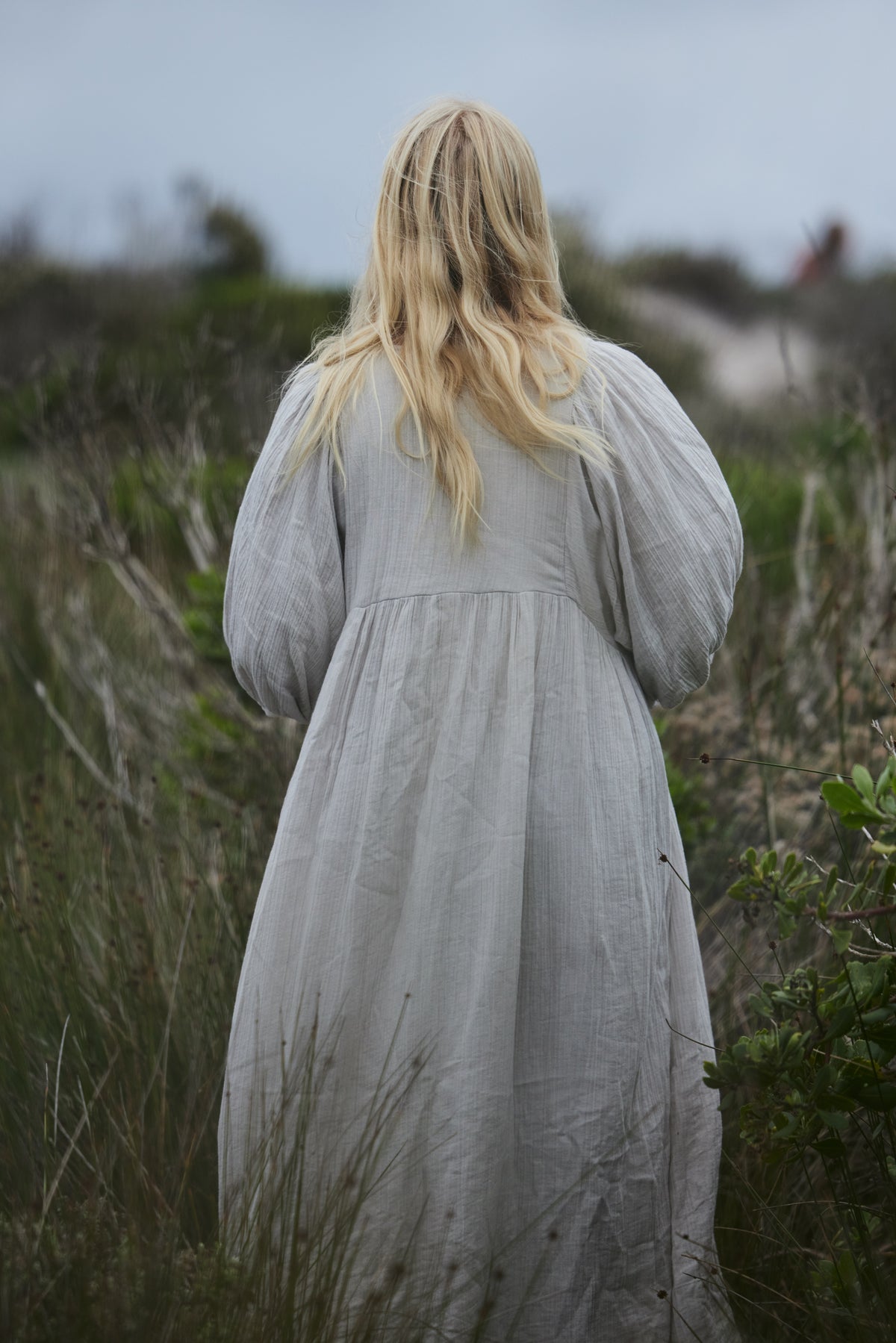 A woman with long blonde hair seen from behind, wearing a long, loose-fitting pale blue organic cotton dress while standing in a grassy outdoor field.