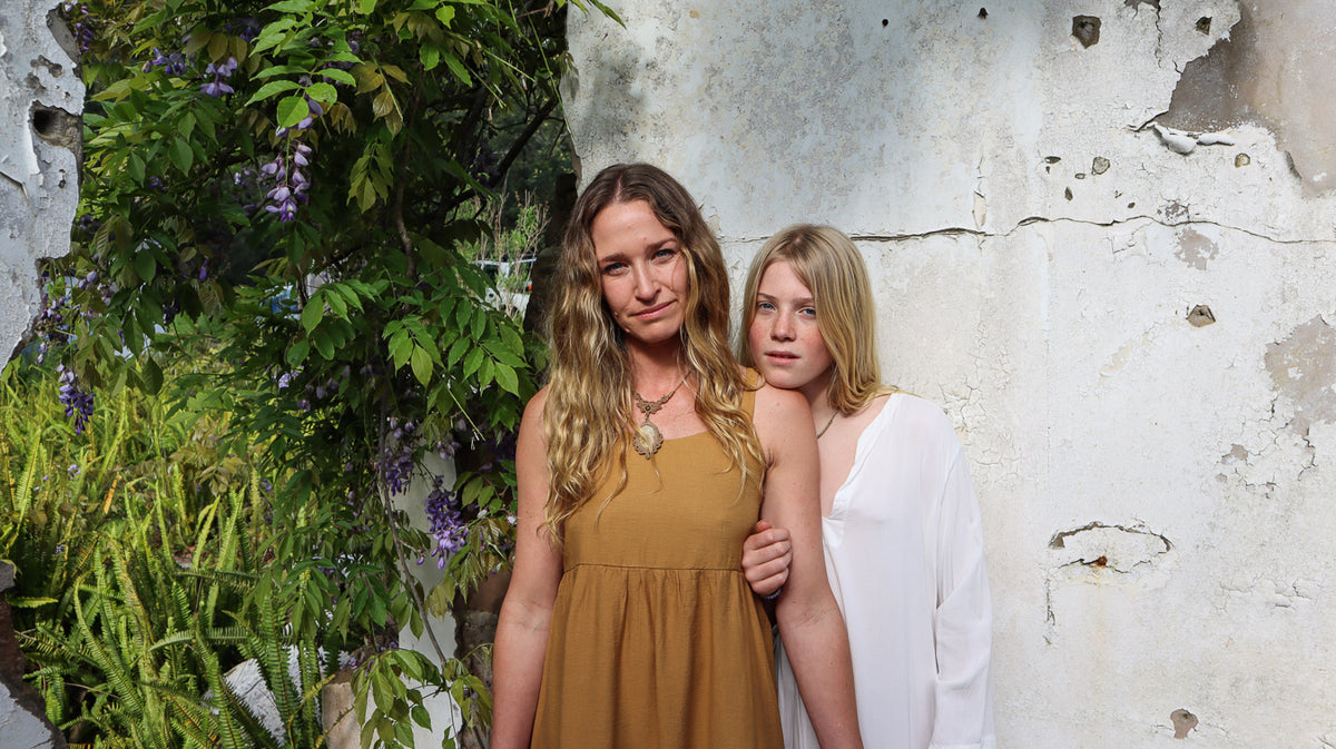 Two young women posing outdoors against a weathered white wall with green foliage on the left. The woman on the left wears a mustard yellow sleeveless dress, and the woman on the right wears a white draped top.