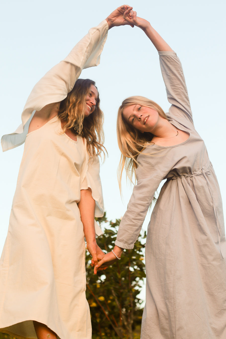 Two women outdoors wearing loose, long-sleeve linen dresses in cream and light taupe, holding hands and stretching their arms upward in a playful pose against a pale sky.