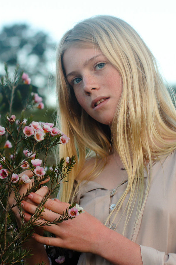 A young blonde woman in a beige top gently holds a pink-flowered bush outdoors, with a softly blurred natural background.
