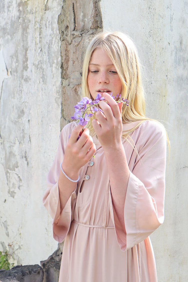 A young blonde woman in a light pink dress holds a small purple flower to her nose, standing against a weathered white and gray stone wall.