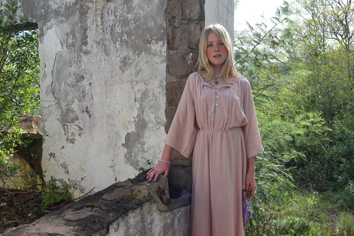 A young blonde woman wearing a dusty pink maxi dress with three-quarter sleeves and a buttoned collar, standing outdoors next to a weathered stone wall with green foliage in the background.