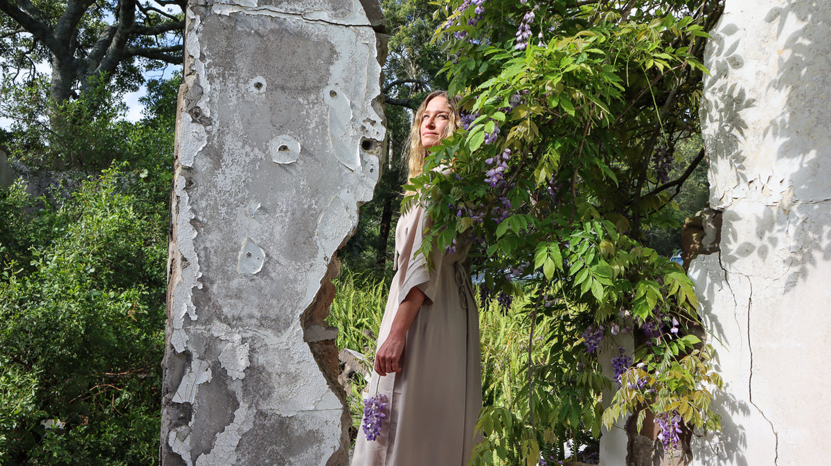 A woman wearing a beige dress stands outdoors between weathered stone pillars, surrounded by lush green foliage and purple wisteria flowers.