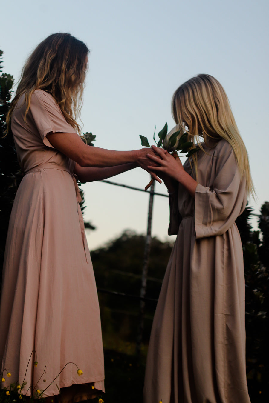 Two women in matching long dusty pink dresses standing outdoors at dusk, holding a white protea between them.