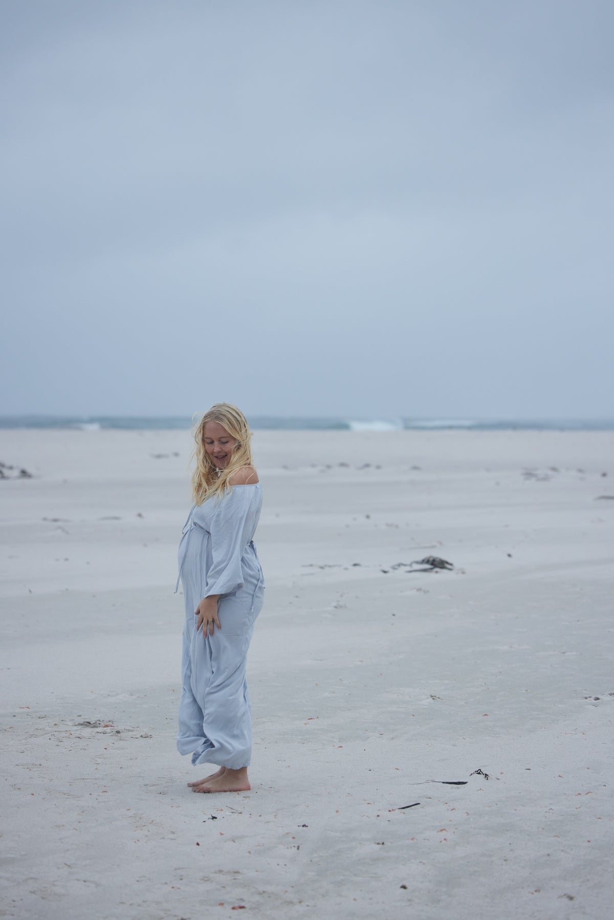 A woman with blonde hair stands barefoot on a wide sandy beach under an overcast sky, wearing a light blue off-the-shoulder long-sleeve jumpsuit.