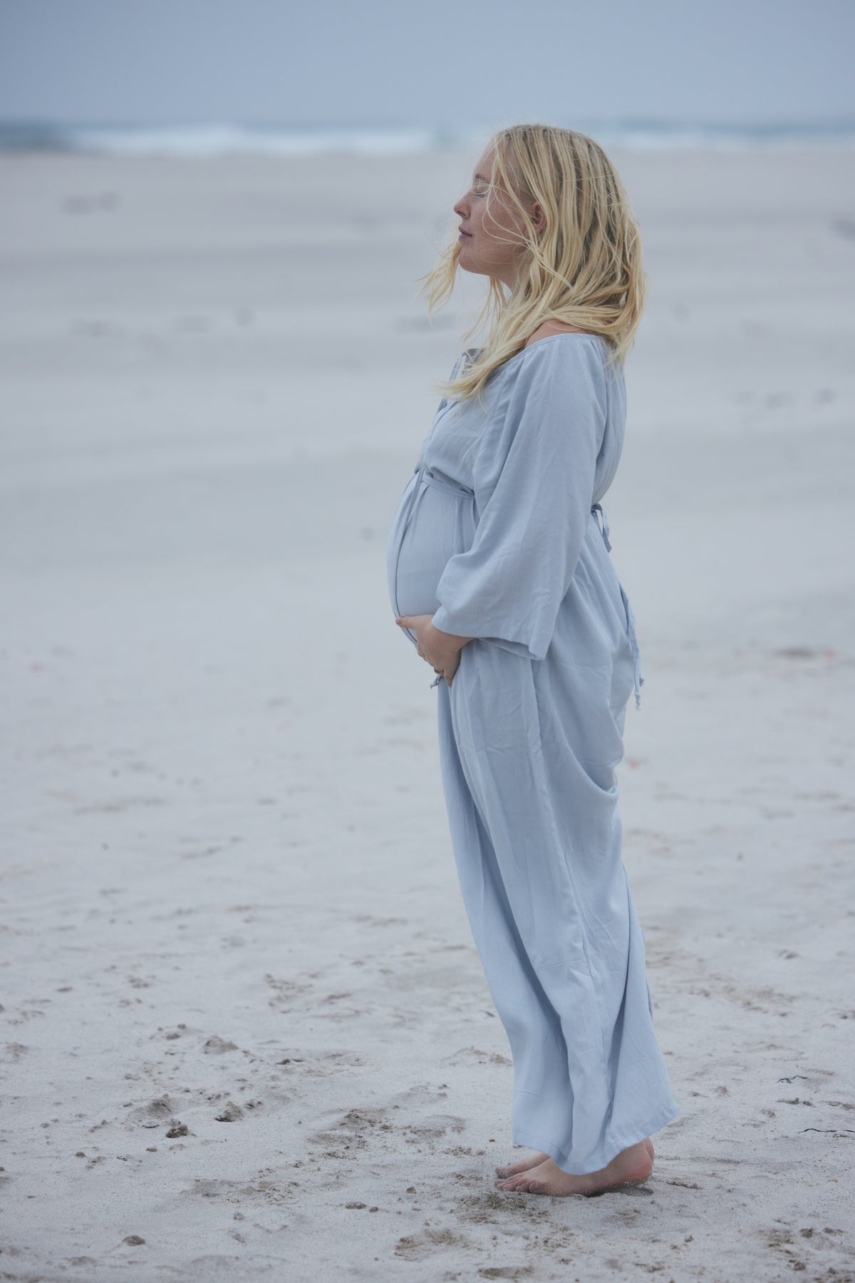 A pregnant woman stands barefoot on a sandy beach wearing a light blue, loose-fitting linen robe or jumpsuit. She is shown in profile, looking up towards the ocean under an overcast sky.