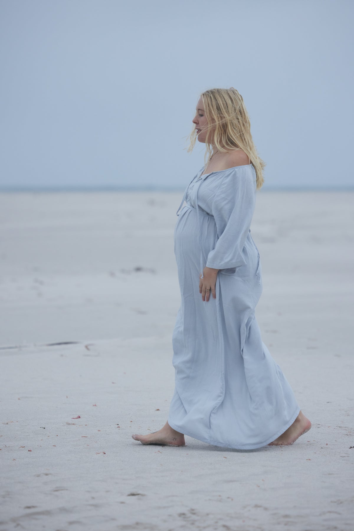 A pregnant woman with blonde hair walks barefoot on a sandy beach wearing a light blue off-the-shoulder maxi dress.