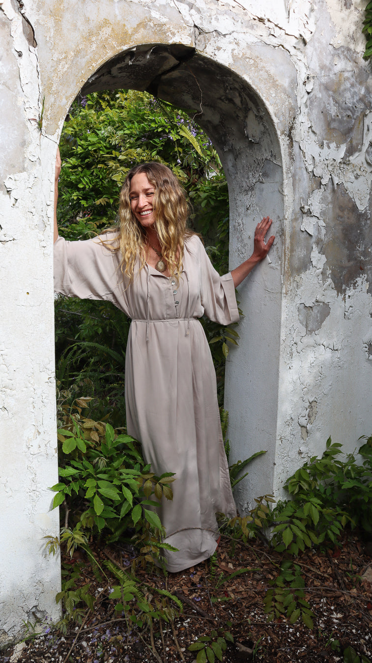 A woman with long blonde hair stands in a white arched stone doorway surrounded by green foliage, wearing a loose, long-sleeved beige maxi dress.