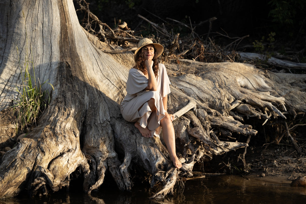 A woman wearing a light beige dress and a wide-brimmed straw hat sits barefoot on the roots of a large tree beside a body of water in a natural outdoor setting.
