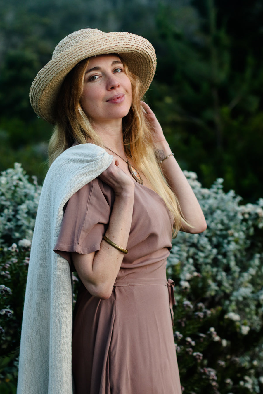 A woman wearing a straw sun hat, a dusty rose-brown dress, and a light cream shawl, posing outdoors in front of a soft-focus background of green foliage and white flowers.