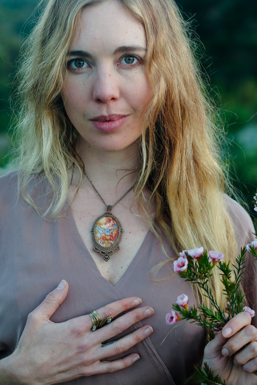 A woman with long blonde hair wearing a bronze pendant necklace with a colorful floral design, holding small pink flowers against a blurred green outdoor background.