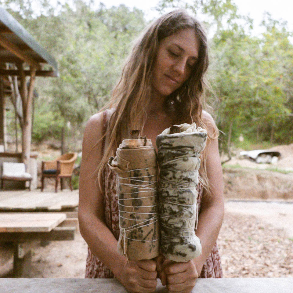 A woman outdoors holding two eco-print wrapped bundles, standing behind a table with trees and a rustic shelter in the background.