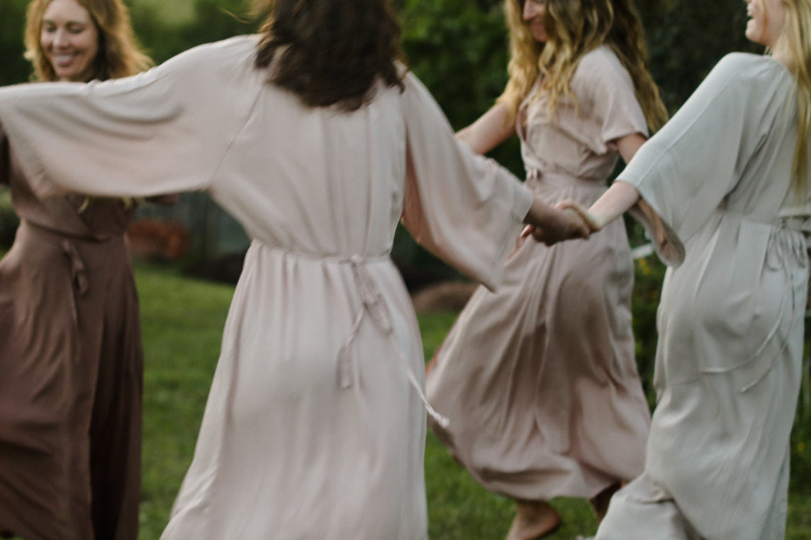 Four women outdoors in a circle wearing flowing maxi-length dresses in soft neutral tones including blush pink, taupe, beige, and stone. The scene is shot from behind the central figure, with grass and trees in the background.