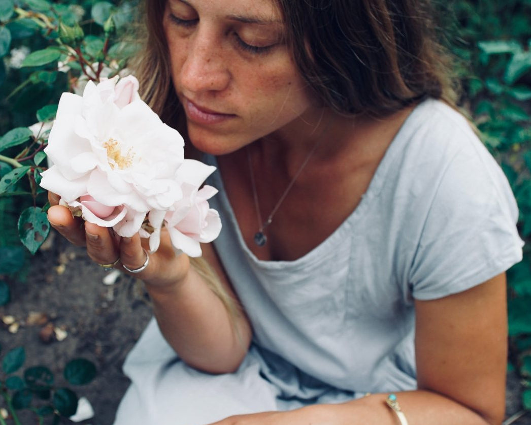 A woman in a light blue dress kneeling in a garden, holding a pale pink rose close to her face.