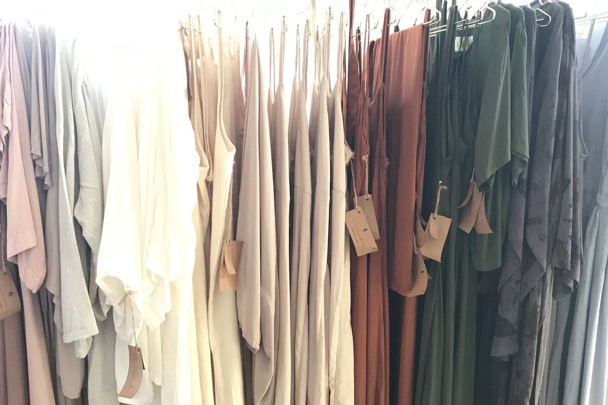A row of hanging garments in soft neutral and earthy tones, including blush, cream, beige, rust, olive green, and charcoal grey, displayed on golden hangers against a bright white background.