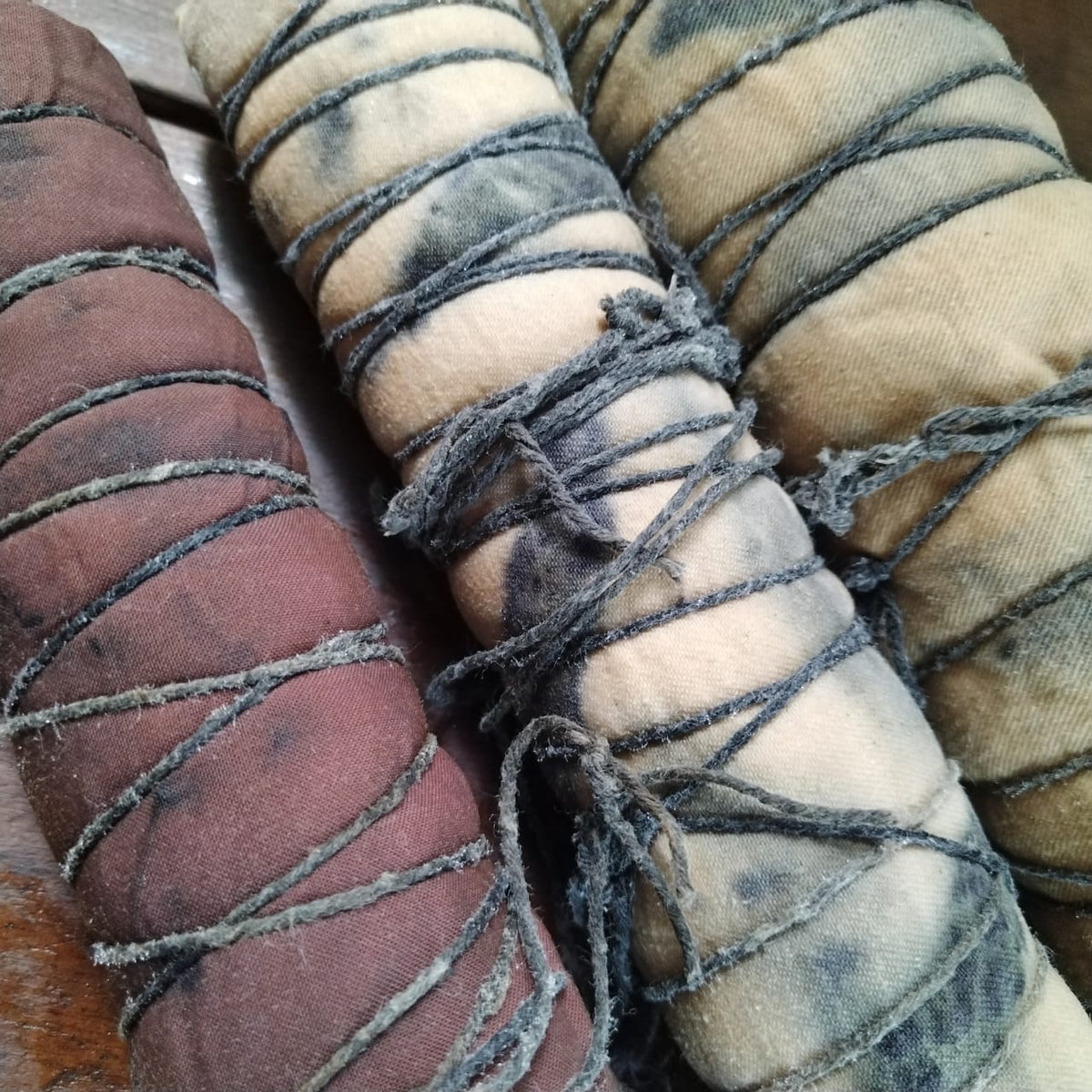 Three hand created eco printed rolls wrapped in dark thread, shown in earthy tones of brown, beige, and grey on a wooden surface.