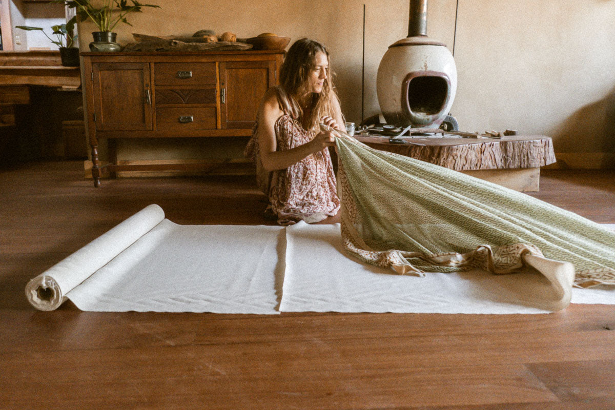 A woman sits on a wooden floor in a rustic room, unrolling a roll of raw silk fabric while holding a light sage green fringed fabric. A wooden cabinet, a white wood-burning stove, and warm wood floors are visible in the background.