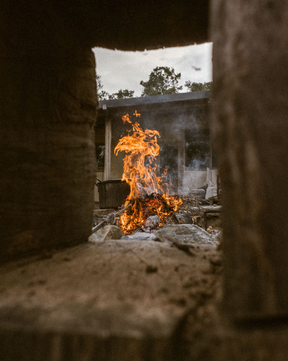 A dramatic fire burning as viewed through a rustic wooden structure, the flames and outdoor scene framed in the centre. 