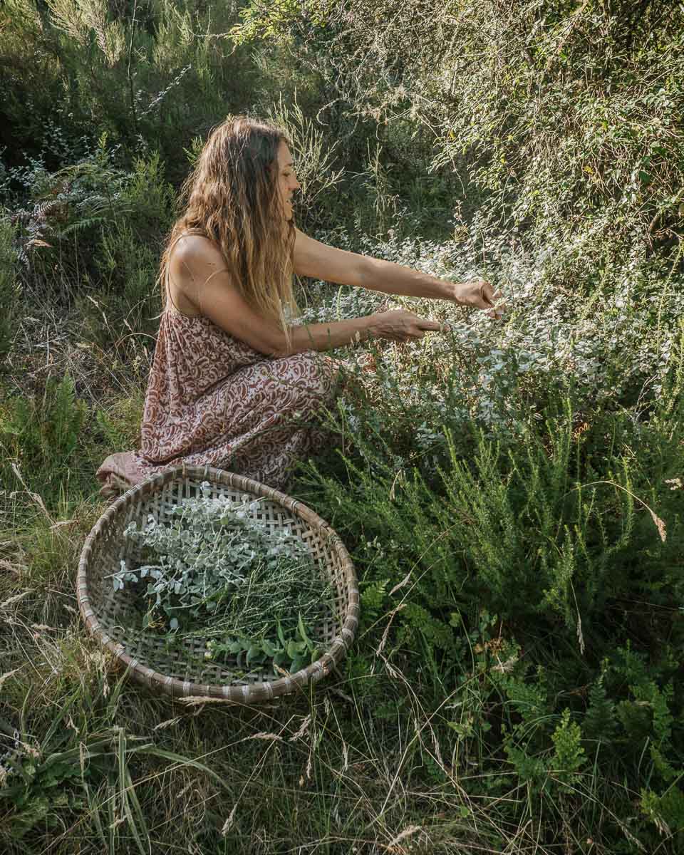 A woman in a brown, patterned dress kneels in a grassy, bushy outdoor setting, reaching toward small white flowers while a woven tray filled with wild botanicals and flowers sits beside her.