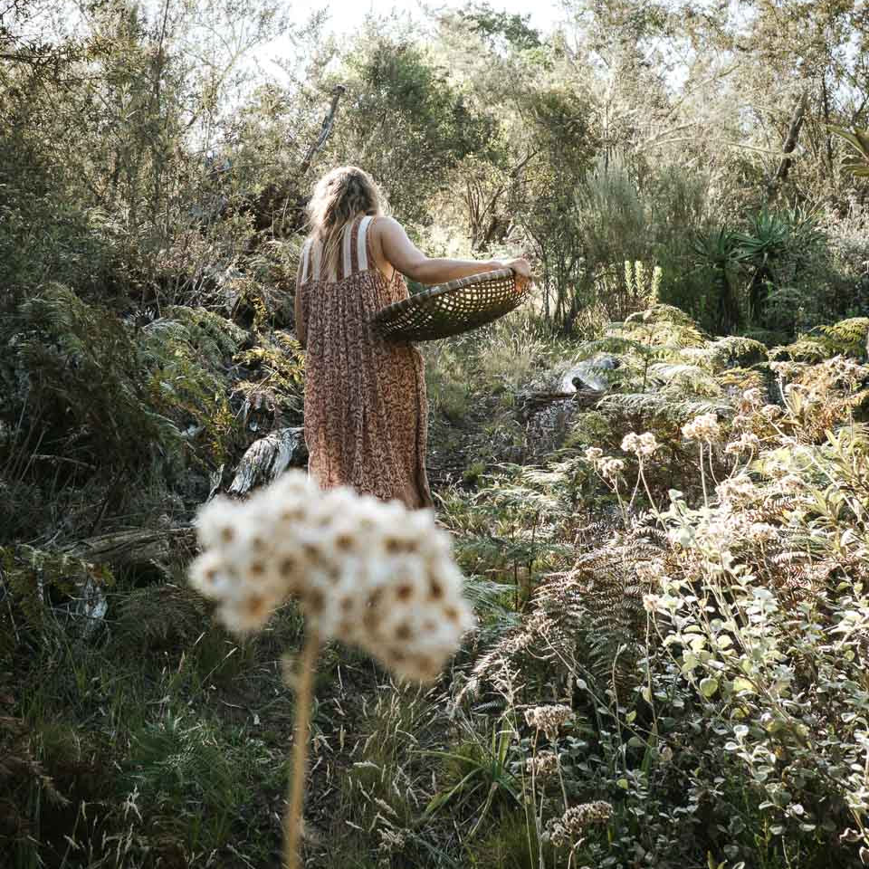 A woman in a brown patterned sleeveless dress stands in a lush, sunlit landscape holding a woven basket, viewed from behind with blurred white flowers in the foreground.