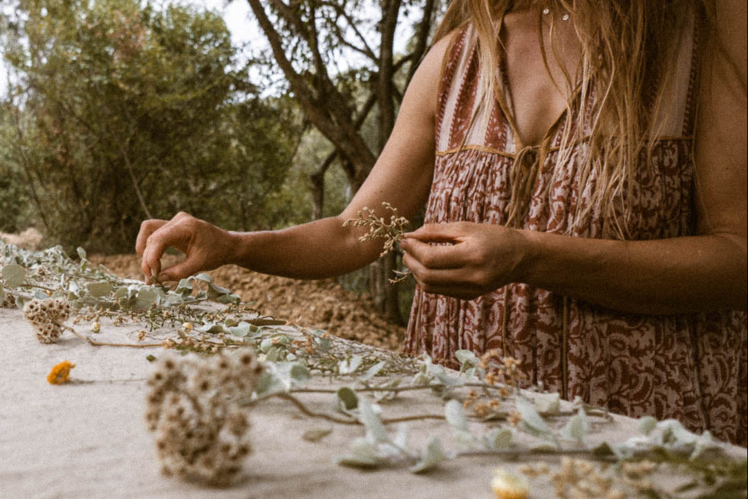 A woman outdoors arranging dried flowers and leaves on a piece of raw silk fabric, wearing a sleeveless rust-brown patterned dress.