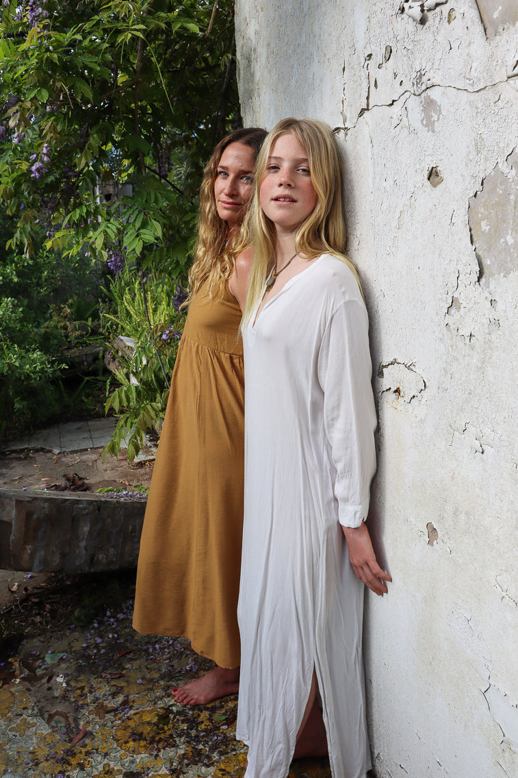 Two women standing outdoors against a weathered white wall, wearing long flowing maxi dresses in white and harvest gold.