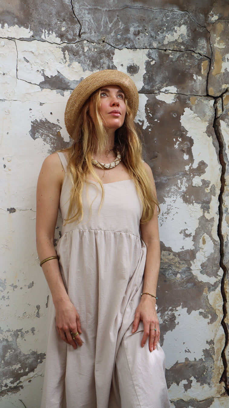 A woman wearing a light beige linen dress and a straw hat stands against a weathered, peeling plaster wall.