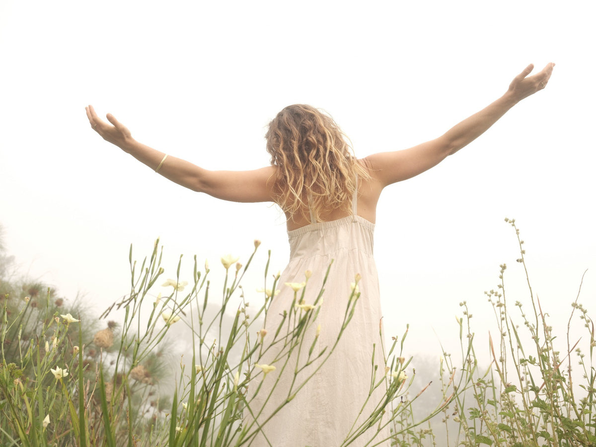 A woman with curly blonde hair stands outdoors in a misty field, viewed from behind with her arms outstretched. She is wearing a flowing white dress, surrounded by tall grasses and small white flowers.