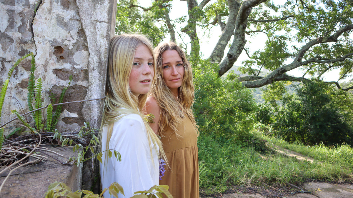 Two women standing outdoors in front of a weathered tree trunk, with lush green foliage and branches in the background. The woman in the foreground wears a white dress, and the woman behind her wears a sleeveless harvest gold dress.