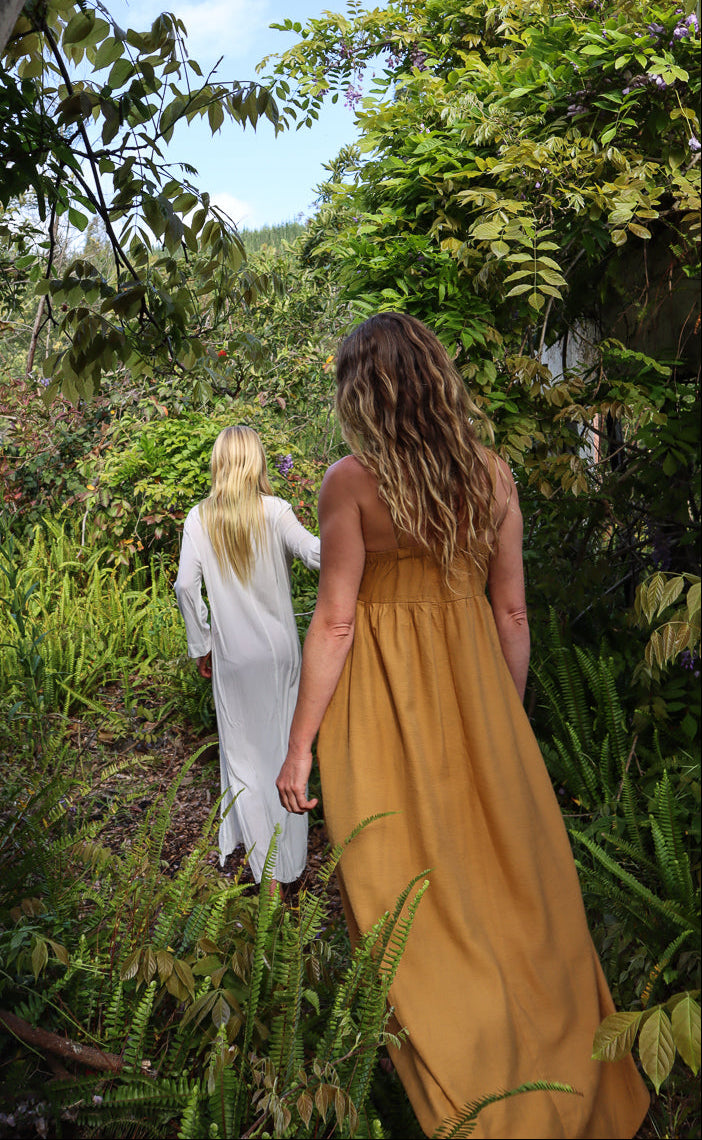 Two women walking outdoors in a garden among pink and white flowers, wearing soft white and harvest gold linen-like dresses.