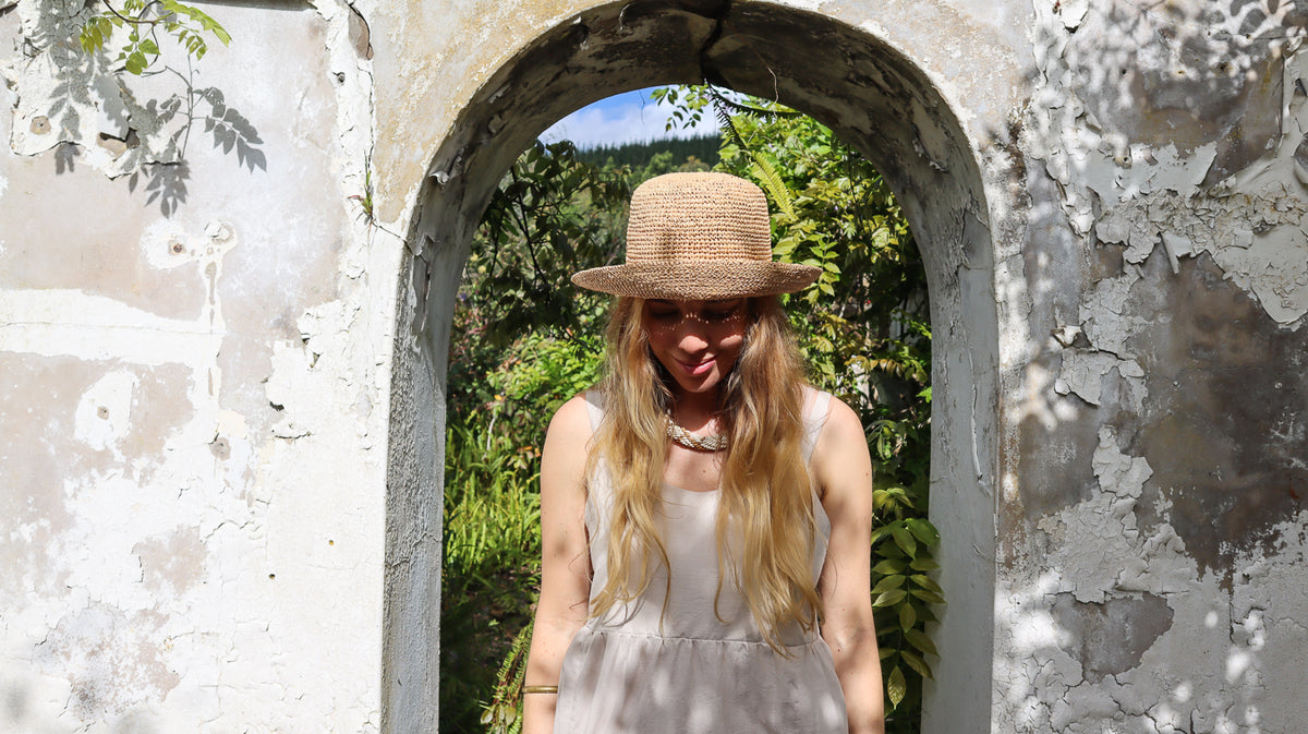 A woman wearing a straw hat, sunglasses, and a light-colored sleeveless dress stands framed by a weathered stone archway with green foliage in the background.