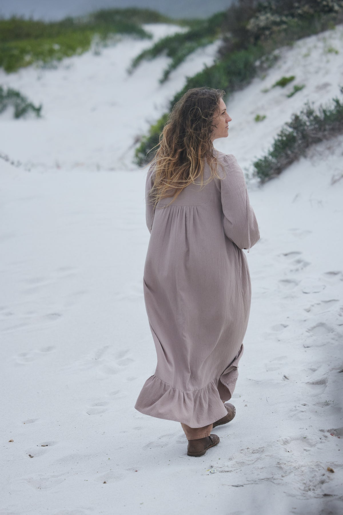 A woman with long wavy light brown hair stands on a white sandy beach, viewed from behind, wearing a long-sleeved dusty mauve maxi dress and brown sandals.