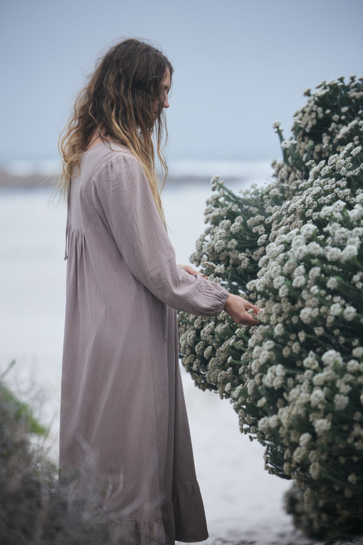 A woman in a long, loose, dusty mauve-pink dress stands outdoors beside a large bush of small white flowers, touching one of the blooms.
