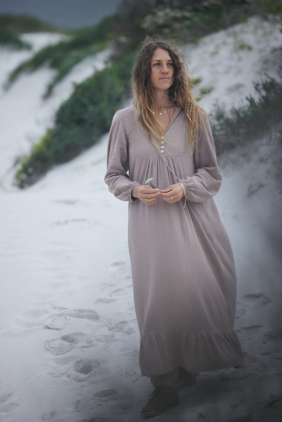 A woman with long, wavy light brown hair stands on a sandy path wearing a long-sleeved, loose-fitting maxi dress in a muted dusty mauve or taupe colour. The dress has a V-neckline with a decorative shell-like button detail at the centre and a gathered, flowing silhouette.