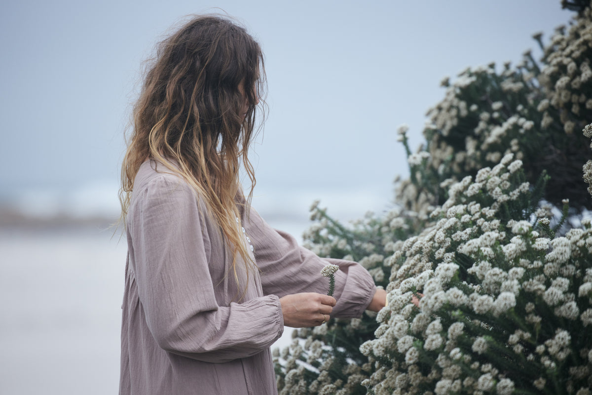 A woman with long, wavy light brown hair wearing a dusty mauve long-sleeve dress stands outdoors next to a bush of small white flowers, reaching toward one of the blooms.